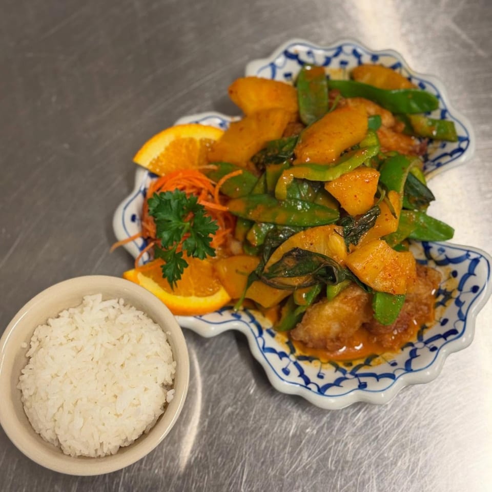 Thai stir-fry with vegetables, orange slices, and a side of white rice on a metal surface.
