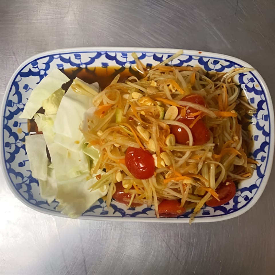 Thai papaya salad with shredded papaya, carrots, tomatoes, peanuts, and cabbage on a blue and white plate.